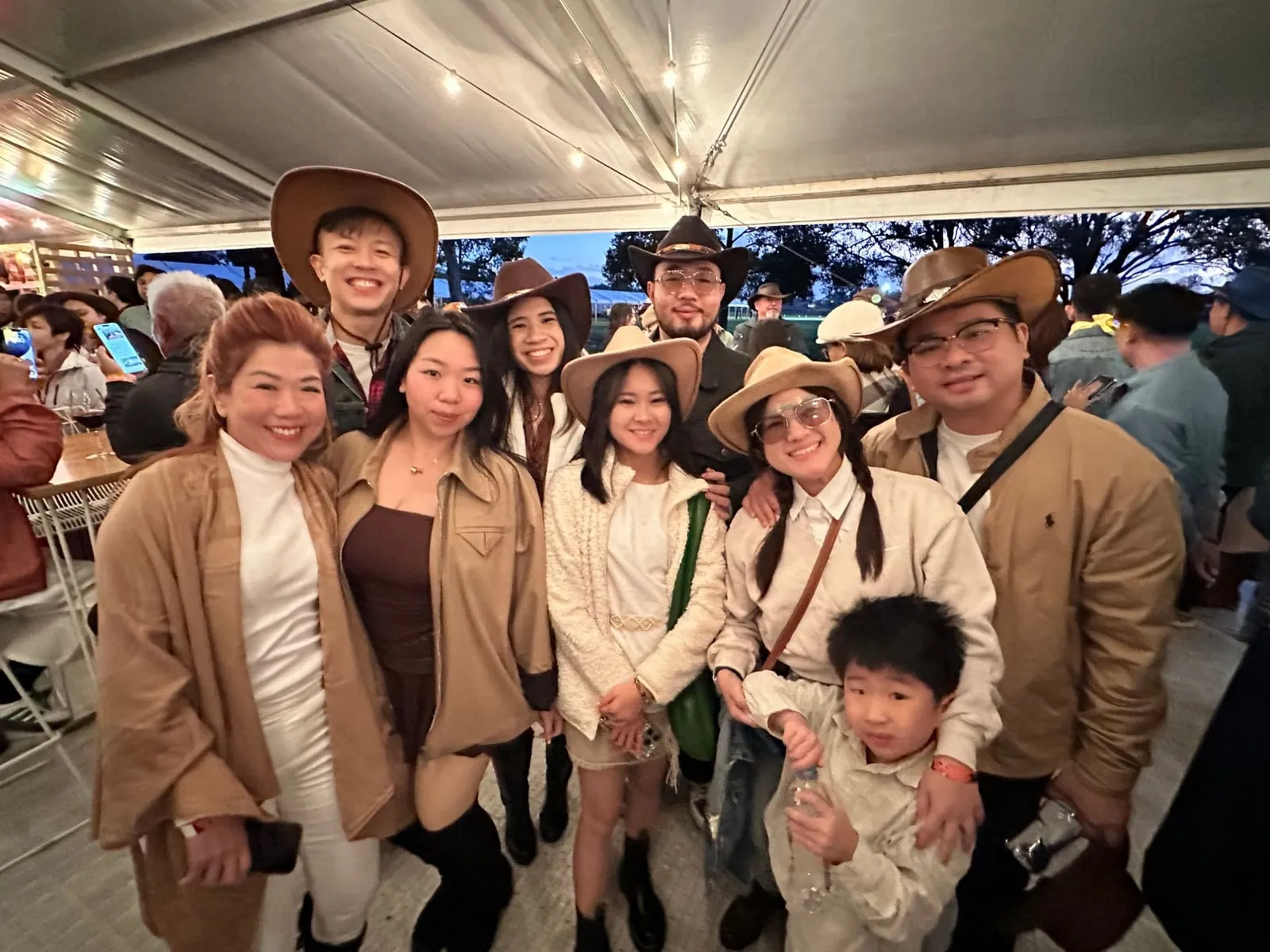 A group in tan and cream outfits, some in cowboy hats, smiling together at an evening event under a tent, with a young boy at the front