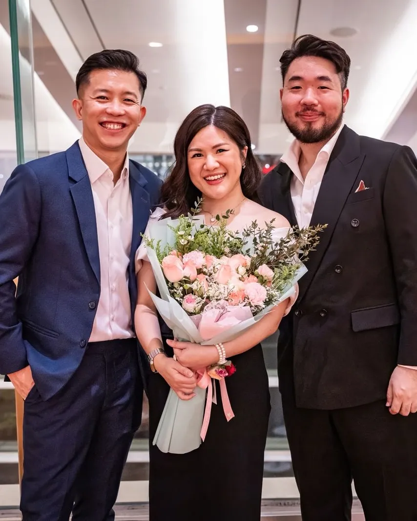 Shermene smiling and holding a bouquet of pink flowers, standing between two men in suits at an evening event