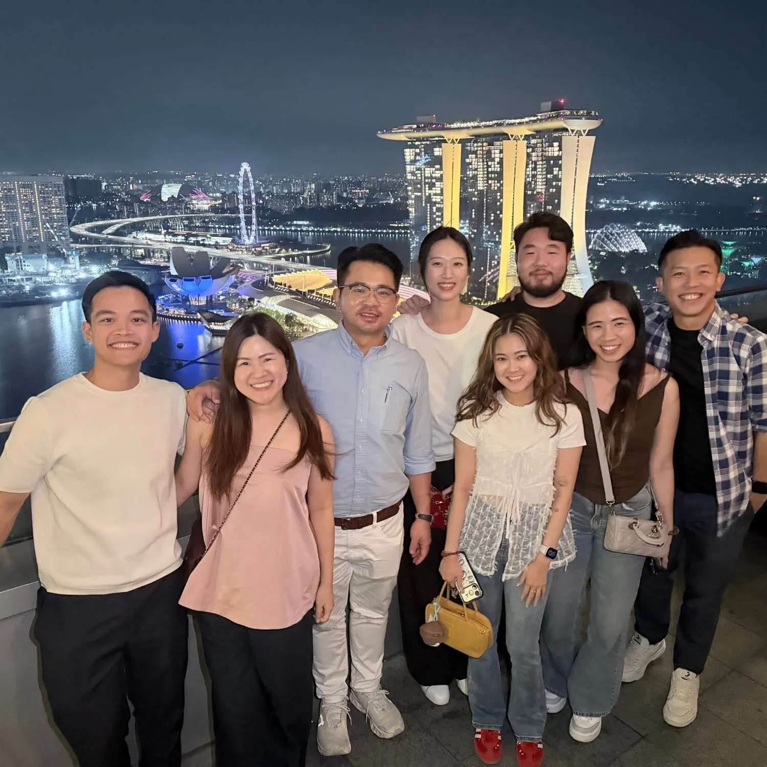 Eight people smiling together on a rooftop at night, with the Marina Bay Sands hotel and the city lights behind them
