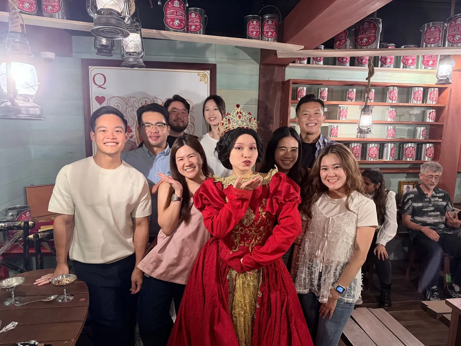 A group photo with a performer dressed as a queen, in front of shelves of red paint tins at a themed event