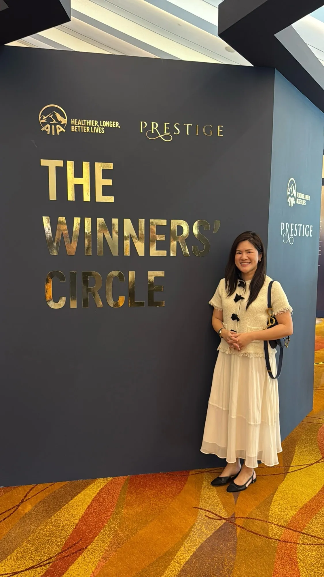 Shermene smiling beside a wall sign that reads The Winners’ Circle, with the AIA and Prestige logos above it